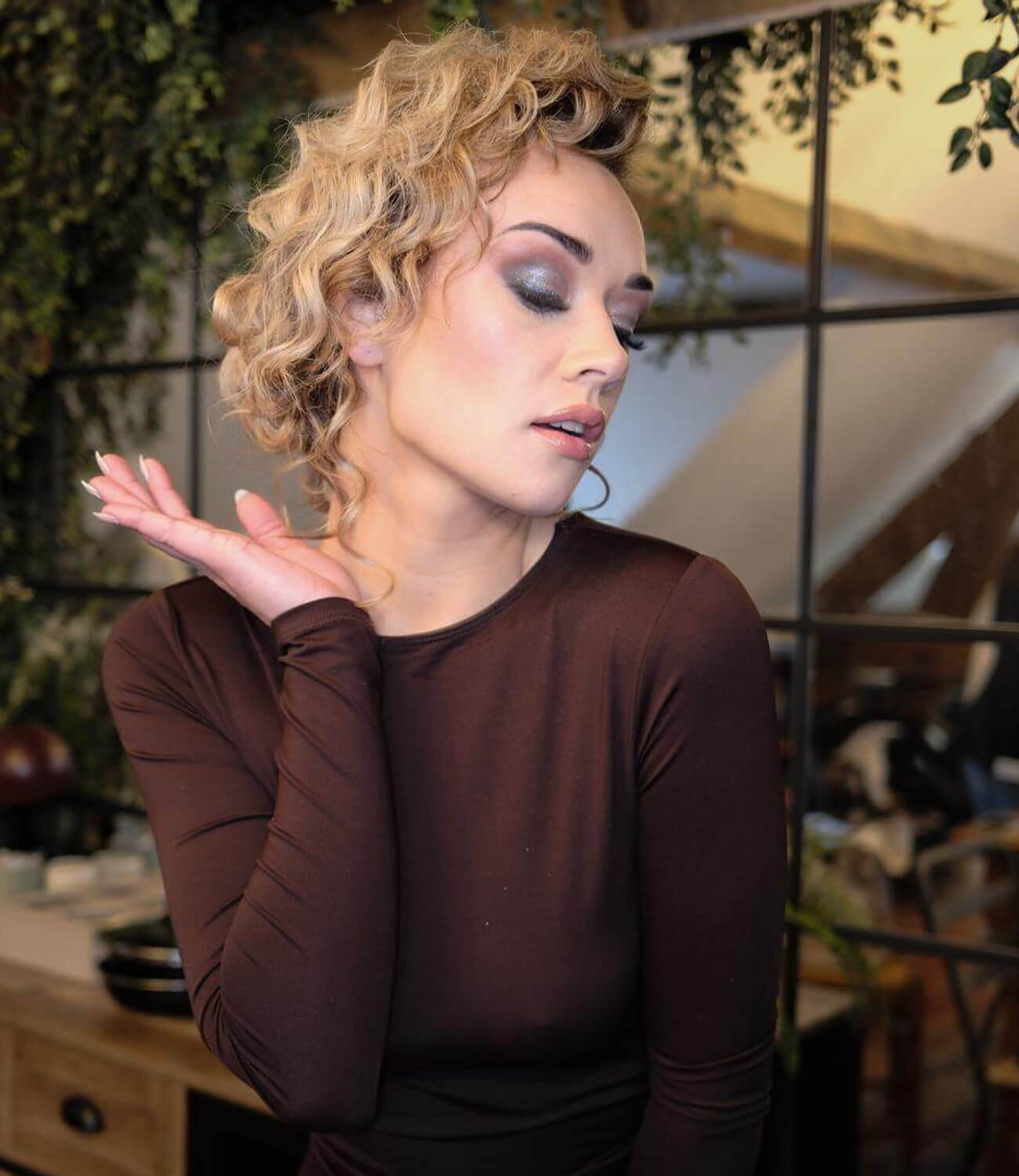 Femme aux cheveux blonds bouclés coiffés en volume vers l’arrière, maquillage scintillant avec paupières fermées, posant avec élégance dans un décor intérieur végétalisé, vêtue d’un top marron à manches longues.