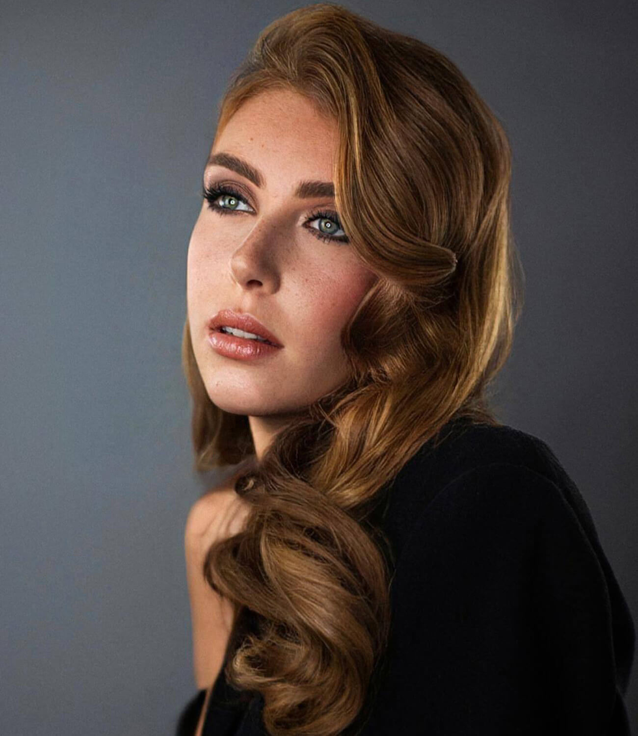 Portrait d’une femme aux cheveux longs cuivrés coiffés en larges ondulations rétro, avec un maquillage glamour et un regard tourné vers le haut, posant sur fond gris en lumière douce.