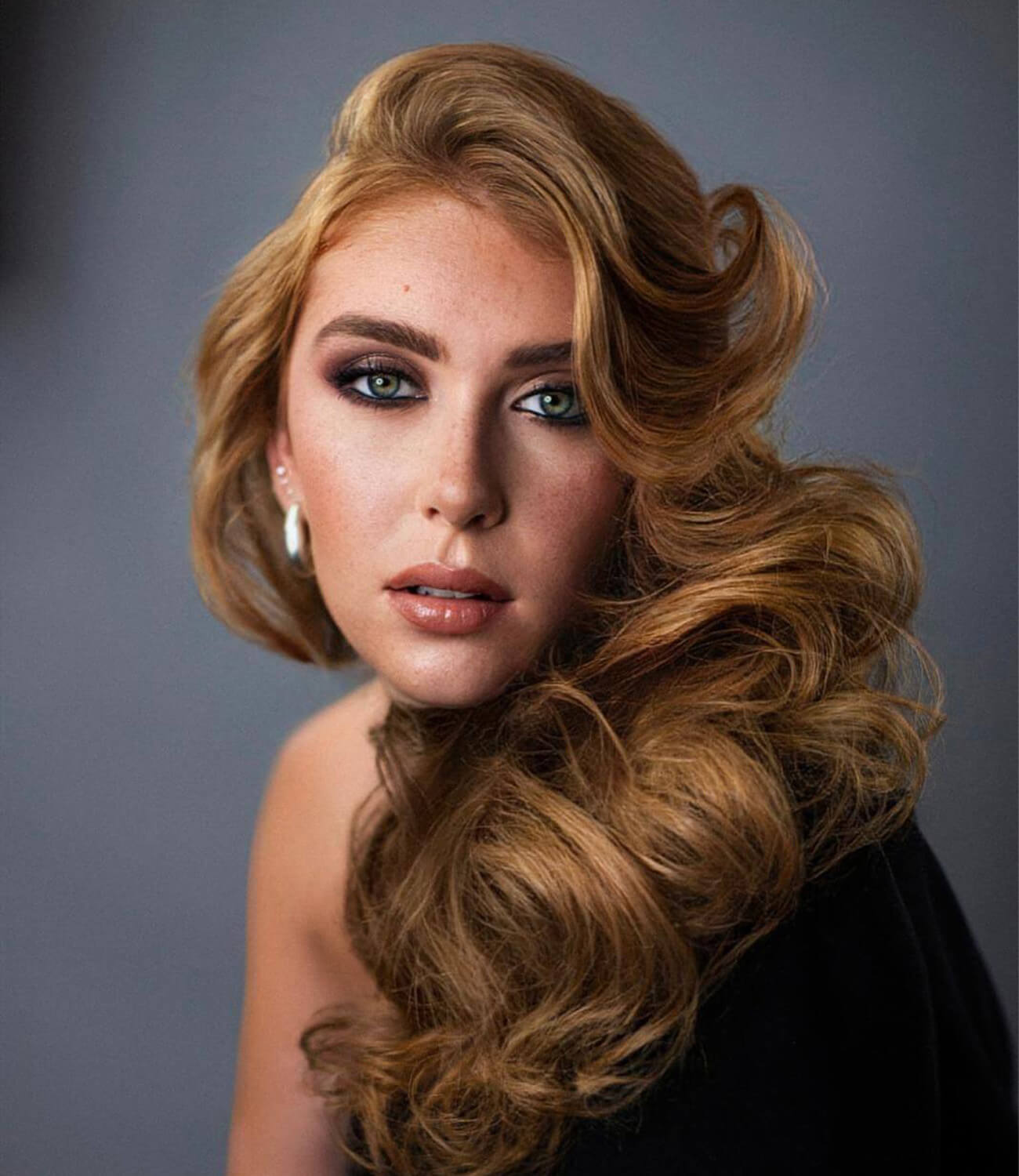 Portrait d’une femme aux cheveux longs, blonds cuivrés et volumineux, coiffés en grandes vagues glamour sur une épaule, avec un maquillage soigné et un regard intense, posant en lumière studio sur fond gris doux.